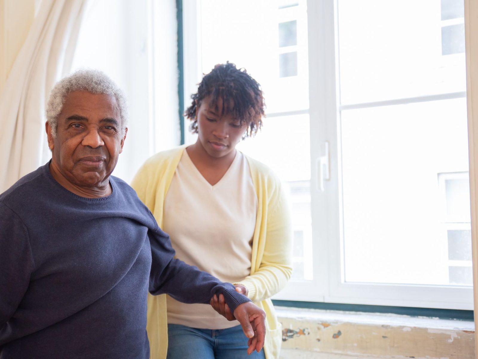 A carer supporting an older man as he walks across a bright room at home