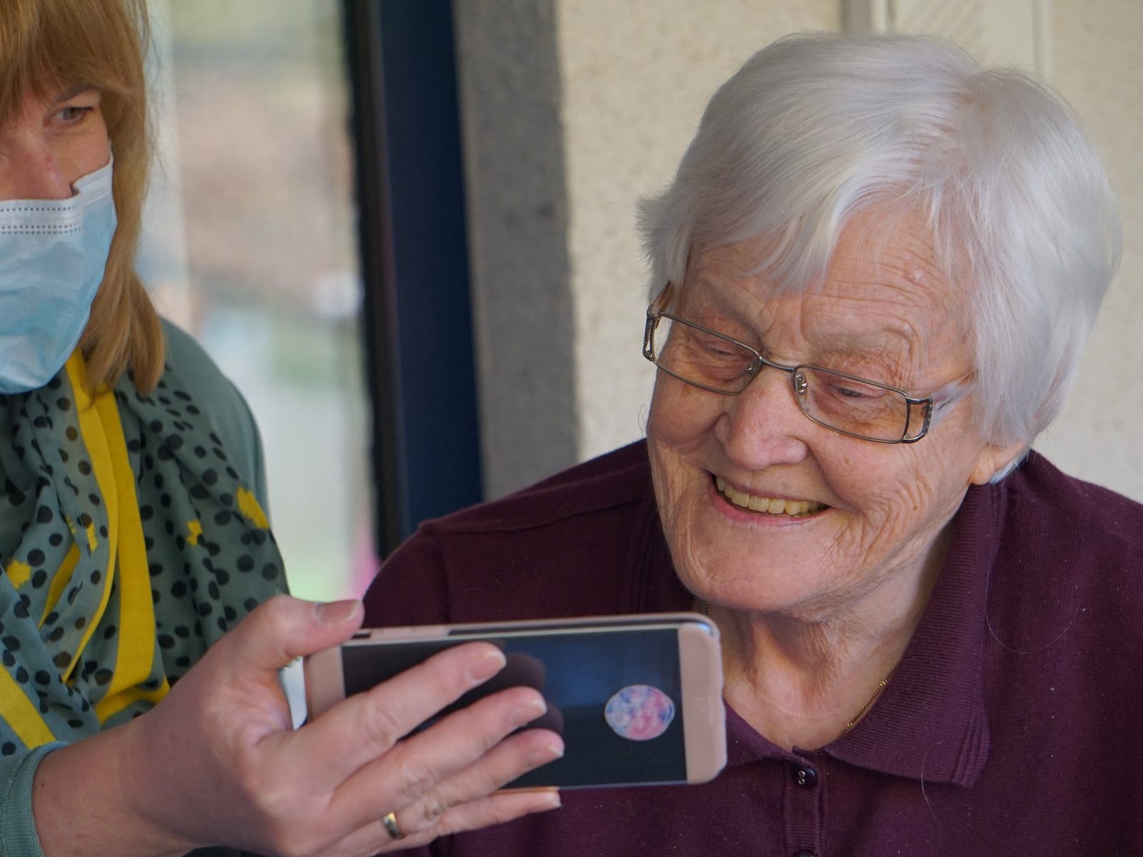 A carer sitting beside an older woman at home, looking at a tablet together